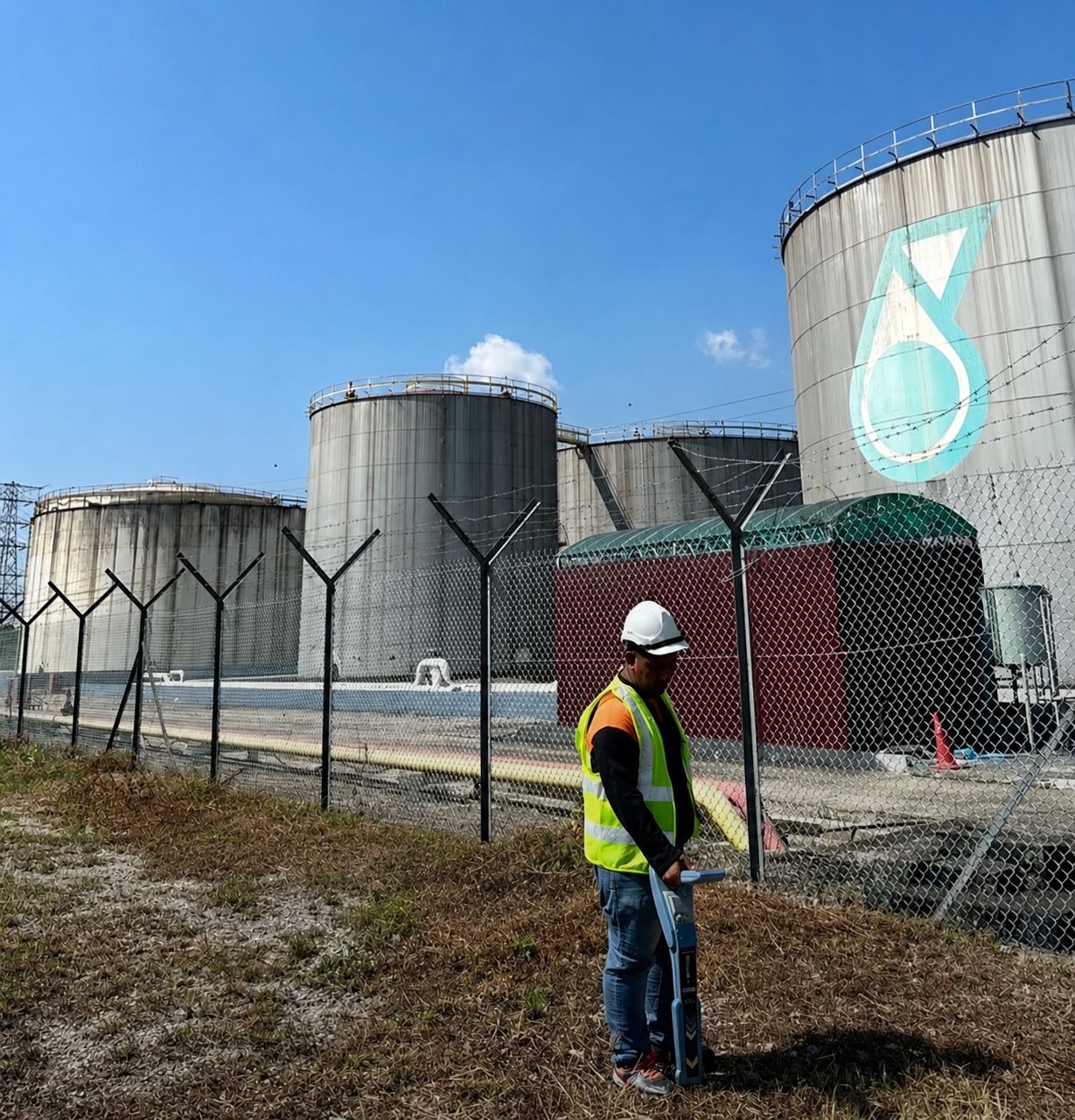 Surveyor tracing underground utilities with an RD8200 locator beside PETRONAS storage tanks